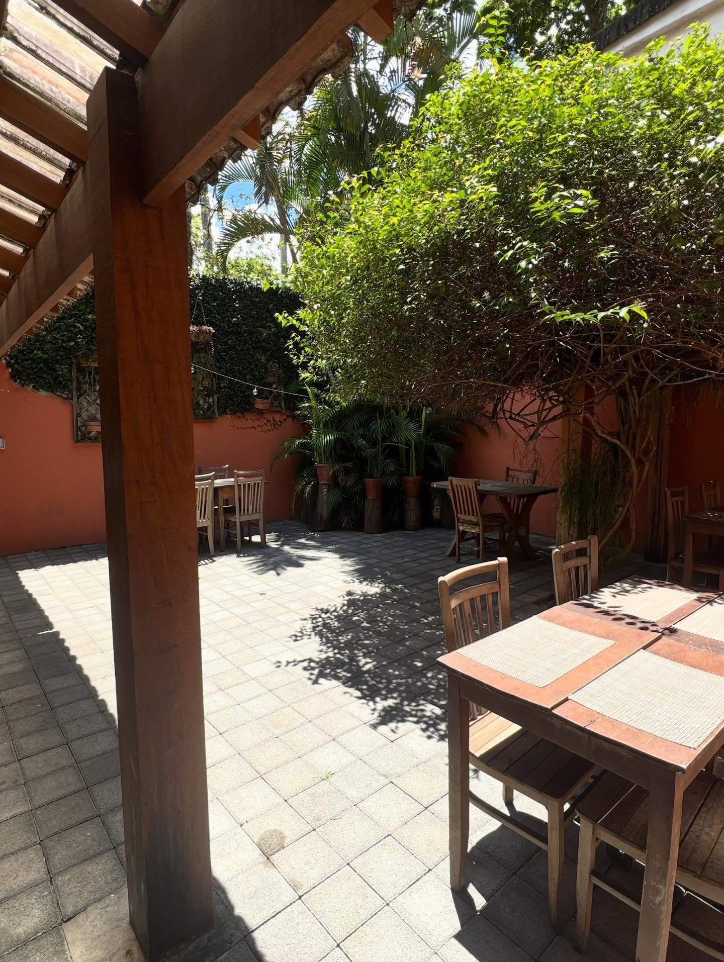 Pátio do Casarão com mesas de madeira sob a pérgola e a árvore ao centro